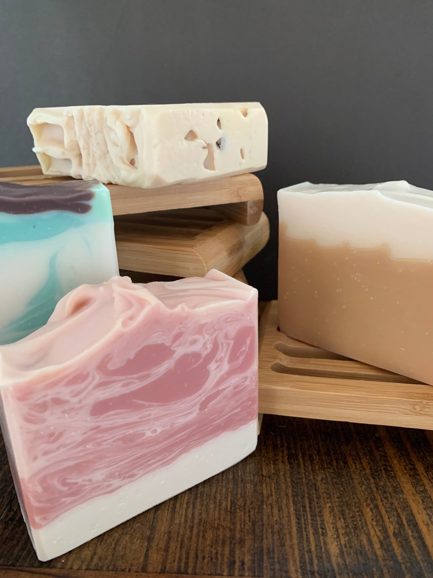 four patisserie inspired fragrant hand made cold press soaps sitting on and around stacked bamboo soap trays. Foreground pink & white raspberry and cream soap, behind is aqua, white and brown mint slice scented soap, back middle is cream coloured cinnamon slice soap & right hand side on bamboo tray sits yellow & cream lemon meringue scented soap.