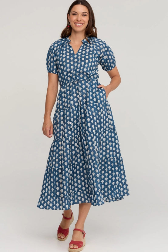 A woman modeling a blue and white patterned midi dress with short puff sleeves, a V-neck, and a self-tie waist, paired with red strappy sandals against a light gray studio background.