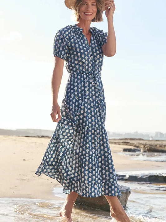 A woman stands barefoot at the edge of the ocean wearing a blue and white patterned midi dress with puff sleeves and a matching hat.