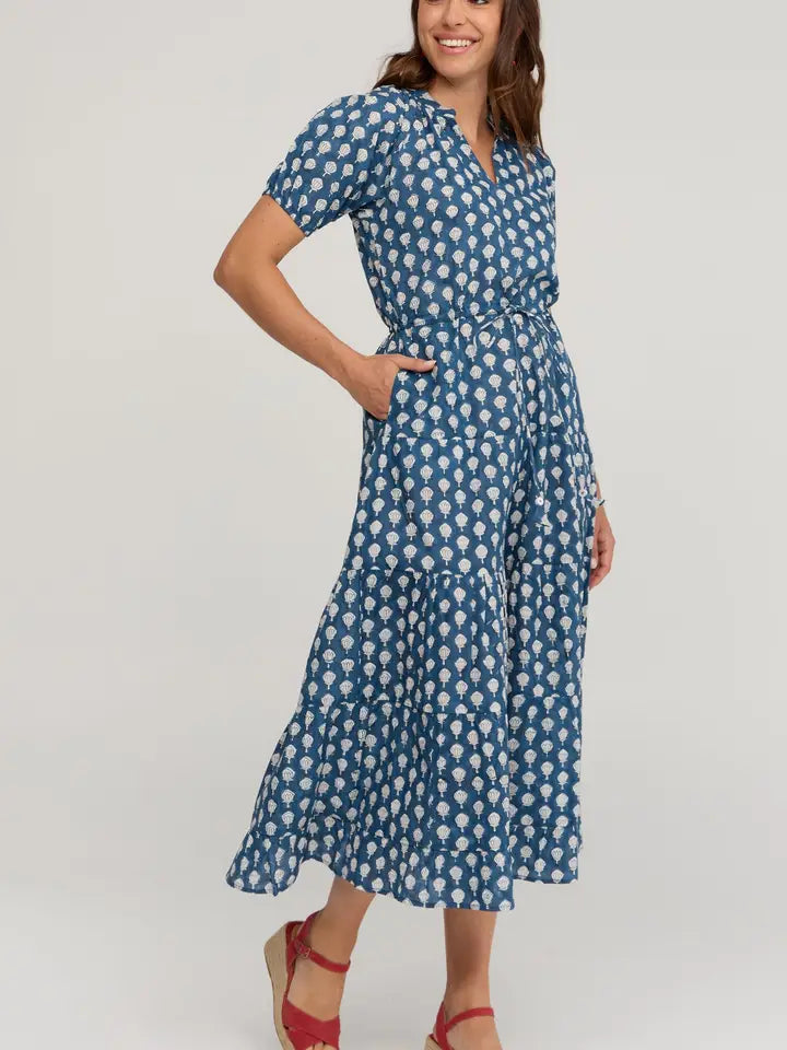 A woman modeling a blue midi dress with a white floral pattern, featuring short sleeves, a V-neckline, and a tie waist, paired with red wedge sandals against a light gray studio background.