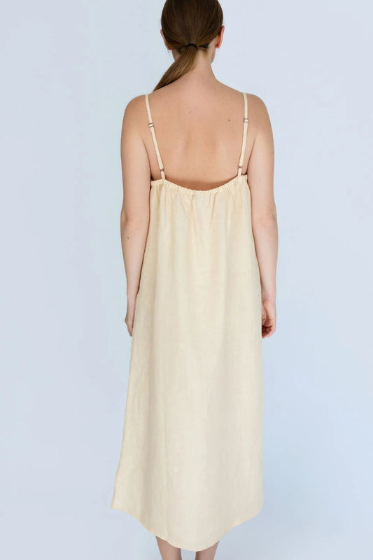 A woman viewed from the back wearing a pale yellow linen slip dress with thin shoulder straps, set against a solid light white background.