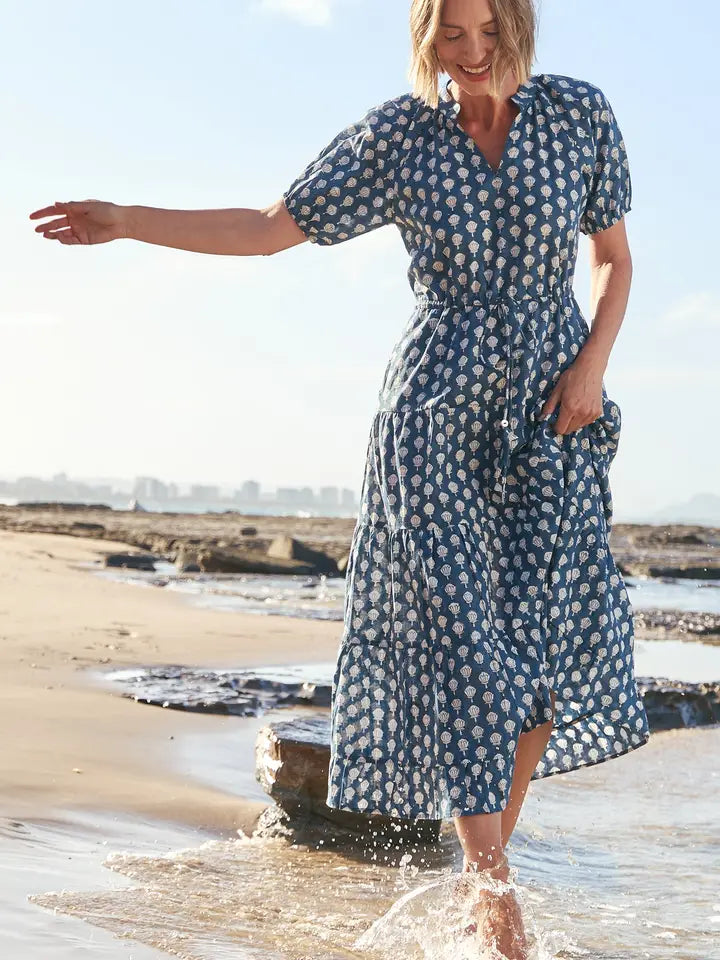 A woman wearing a blue and white patterned midi dress walks through shallow ocean water on a sandy beach.