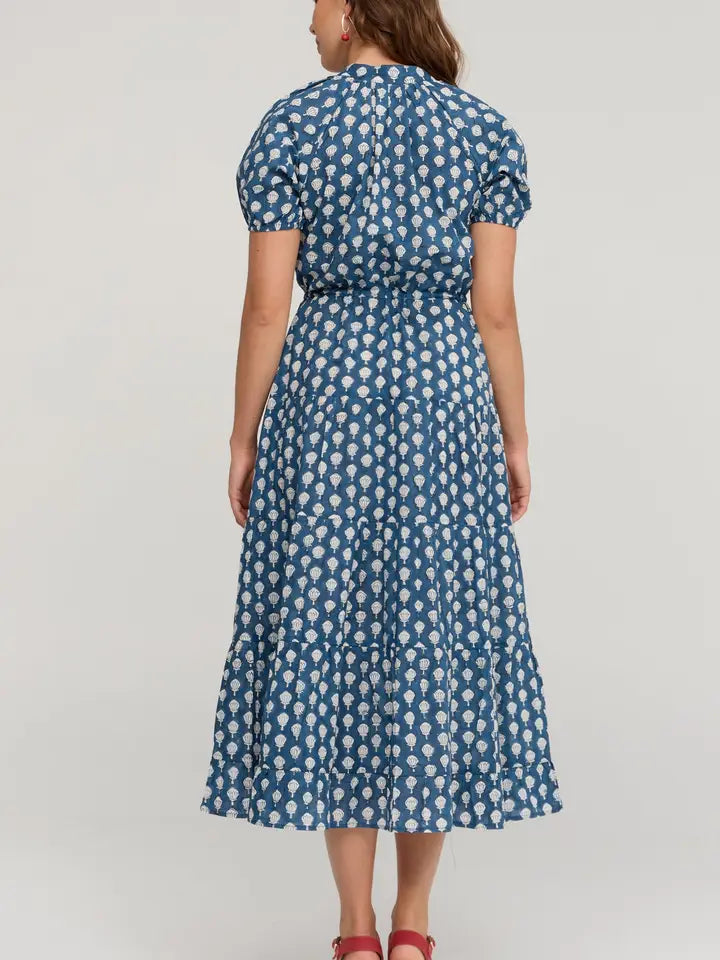 A woman viewed from the back wearing a blue midi dress with a white floral block print pattern, puff sleeves, and a tie at the back neckline, paired with red shoes against a light gray studio background.